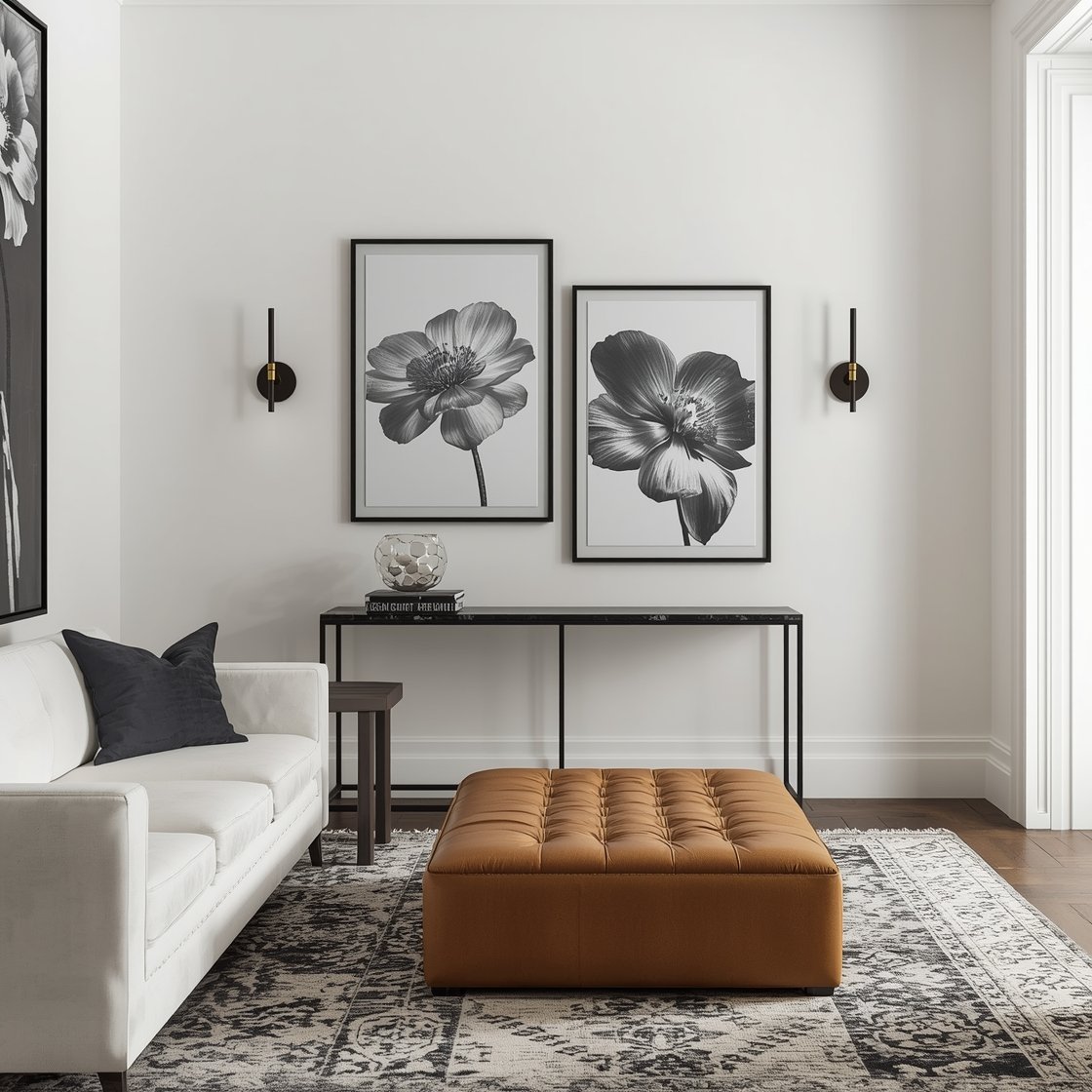 Medium shot, corner angle: A crisp Parisian-influenced living room vignette with plaster-white walls and strong matte black accents. Slender black metal étagères holding stacked art grids and oversized black-and-white floral prints; a slimline black marble console below. In front, a plush white slipcovered sofa faces a tufted camel leather ottoman used as a table, with a charcoal-and-ivory Turkish rug underfoot. Sculptural antique brass sconces flank architectural molding. Finishes include blackened steel, honed marble, and brass; accent tones of charcoal, ecru, and ink. Photorealistic, bright balanced daylight with slight contrast to emphasize black elements.