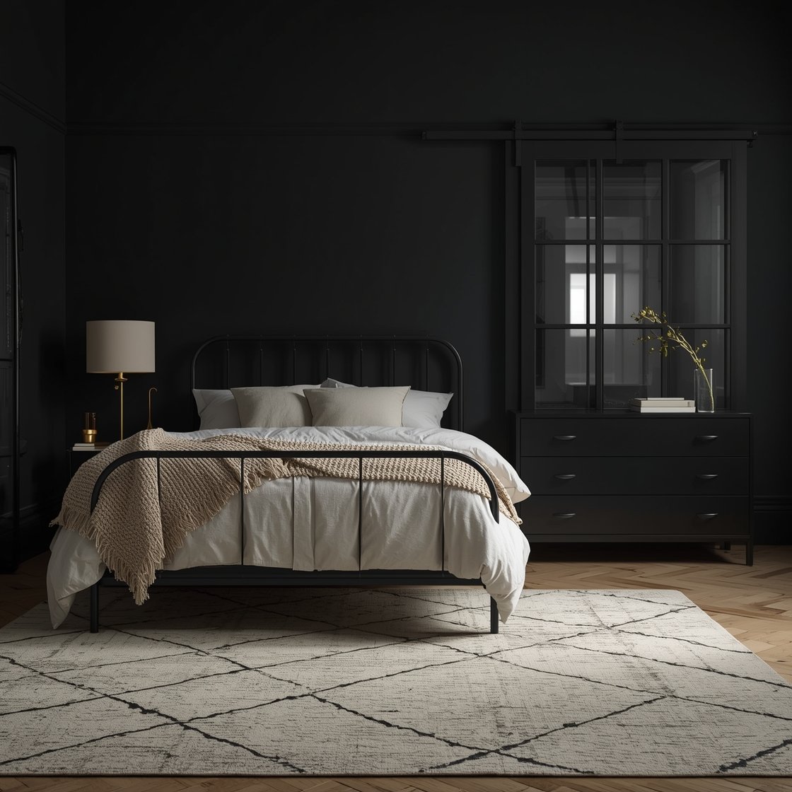Medium shot — Soft Industrial Noir: Charcoal-black walls set a moody backdrop for a cream bedscape—fluffy duvet, oversized euro shams, and a textured knit throw. A black metal bed with curved spindle headboard, cream drum bedside lamps, and a matte black locker-style dresser with soft-close drawers. Light oak floor, large cream Moroccan-style rug with subtle diamond pattern, and a black sliding barn door with frosted glass partially visible. Low, cozy evening lighting.