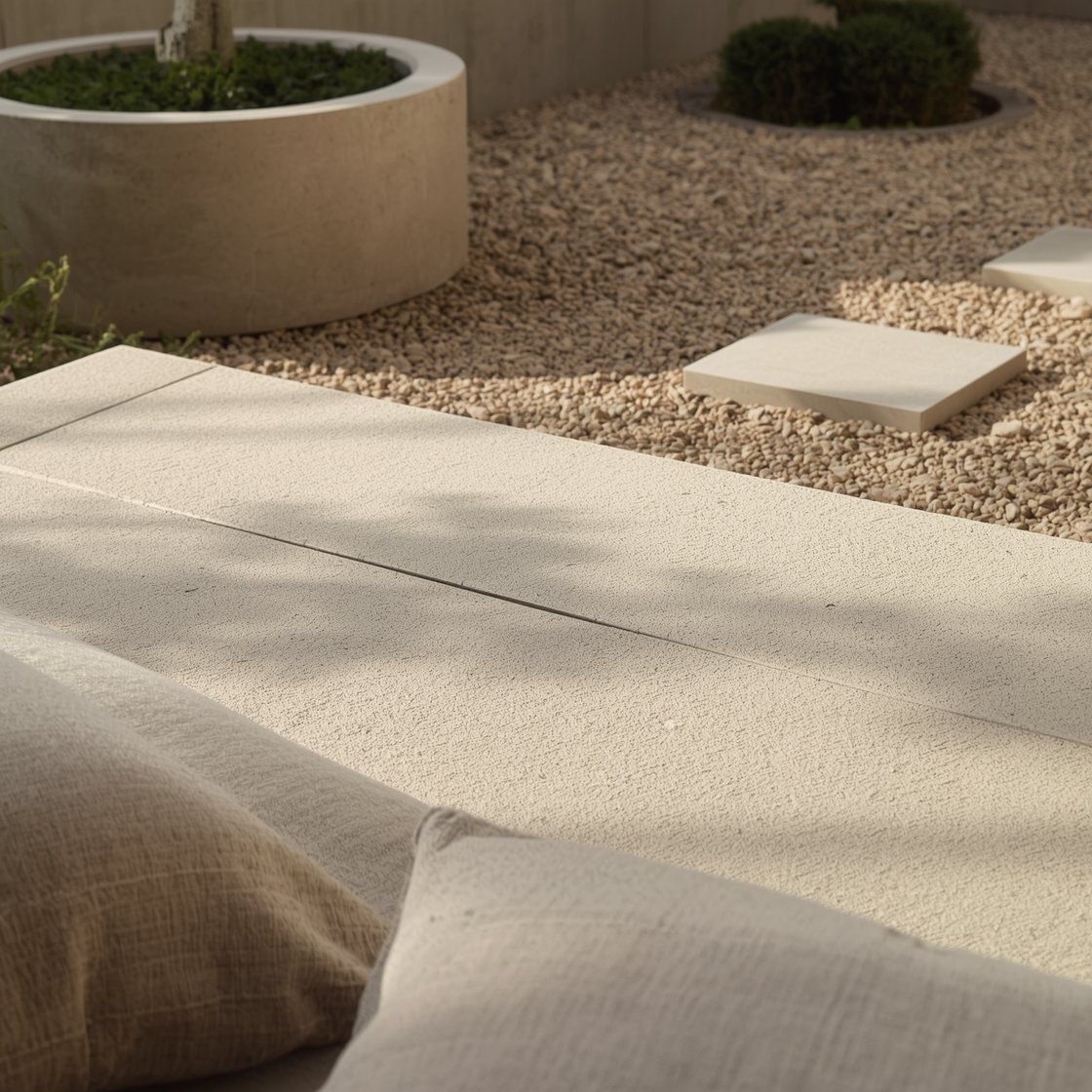 Detail closeup, Japandi serenity textures in soft morning light: sand-finished concrete slab surface with silky, non-shiny feel and calm grid of expansion joints; edge of a slatted cedar platform bench meeting a smooth concrete bowl fire pit; pale ceramic cylinder planter with dwarf pine and moss at the base; linen parasol fabric and neutral floor cushions partially in frame; stepping stones set into pea gravel transitioning to garden beyond; tones of bone, warm sand, soft taupe, and moss; minimal, tactile, serene mood, no people.